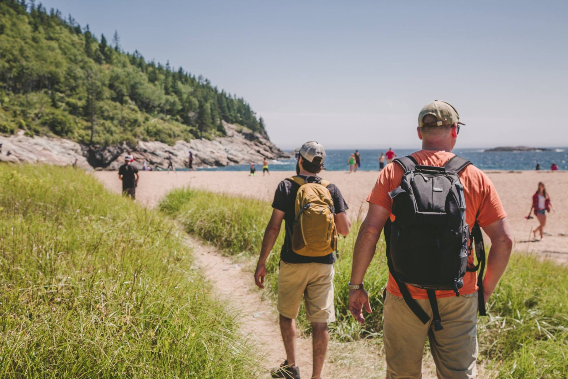 Two adults with backpacks walking a sandy path through beach grass toward the shore