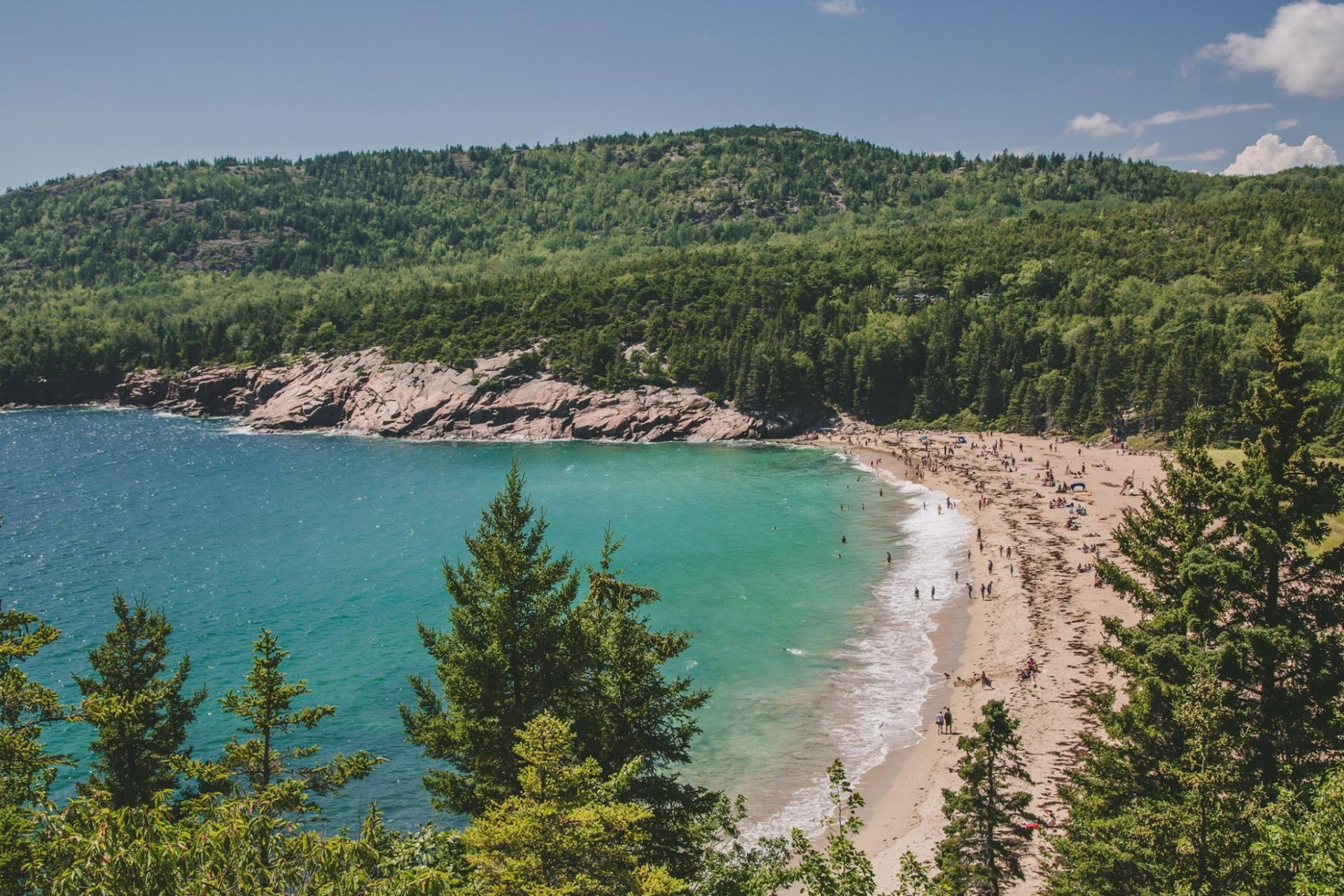 Sand Beach in Acadia National Park seen from a wooded overlook, turquoise water and pink granite cliffs
